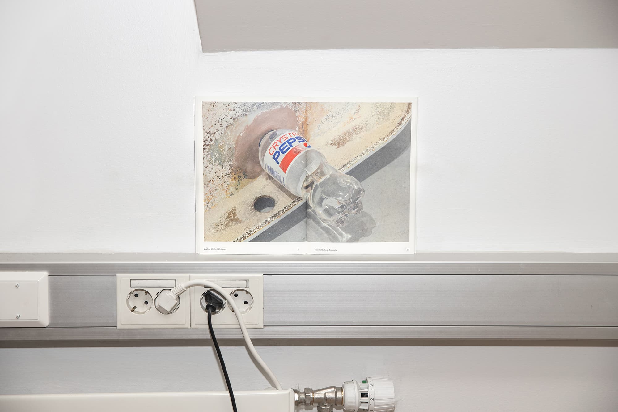 A folded out book sits on a chrome sill in an office. The book spread shows a detail of an artwork. A crystal pepsi bottle is attached to a bathtub.