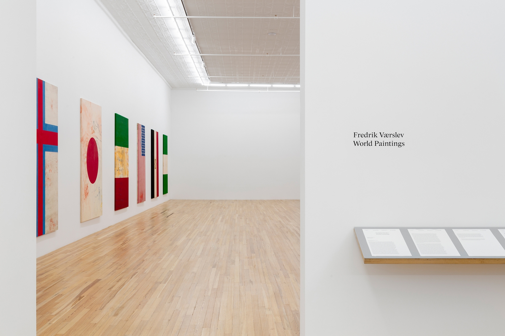 Exhibition text in a gallery. Six paintings og flags hang on the left side wall. Exhibition material sits on an aluminum shelf below vinyl text.
