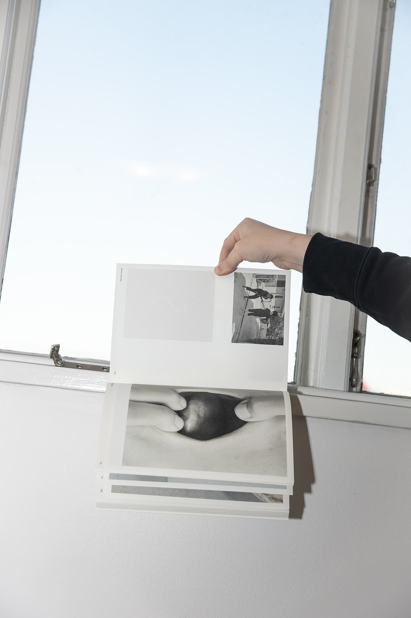 Someone in a black sweater holds open a book in a window sill. Two black and white images are visible on the spread. The light from the window makes the image on the back of the page visible.