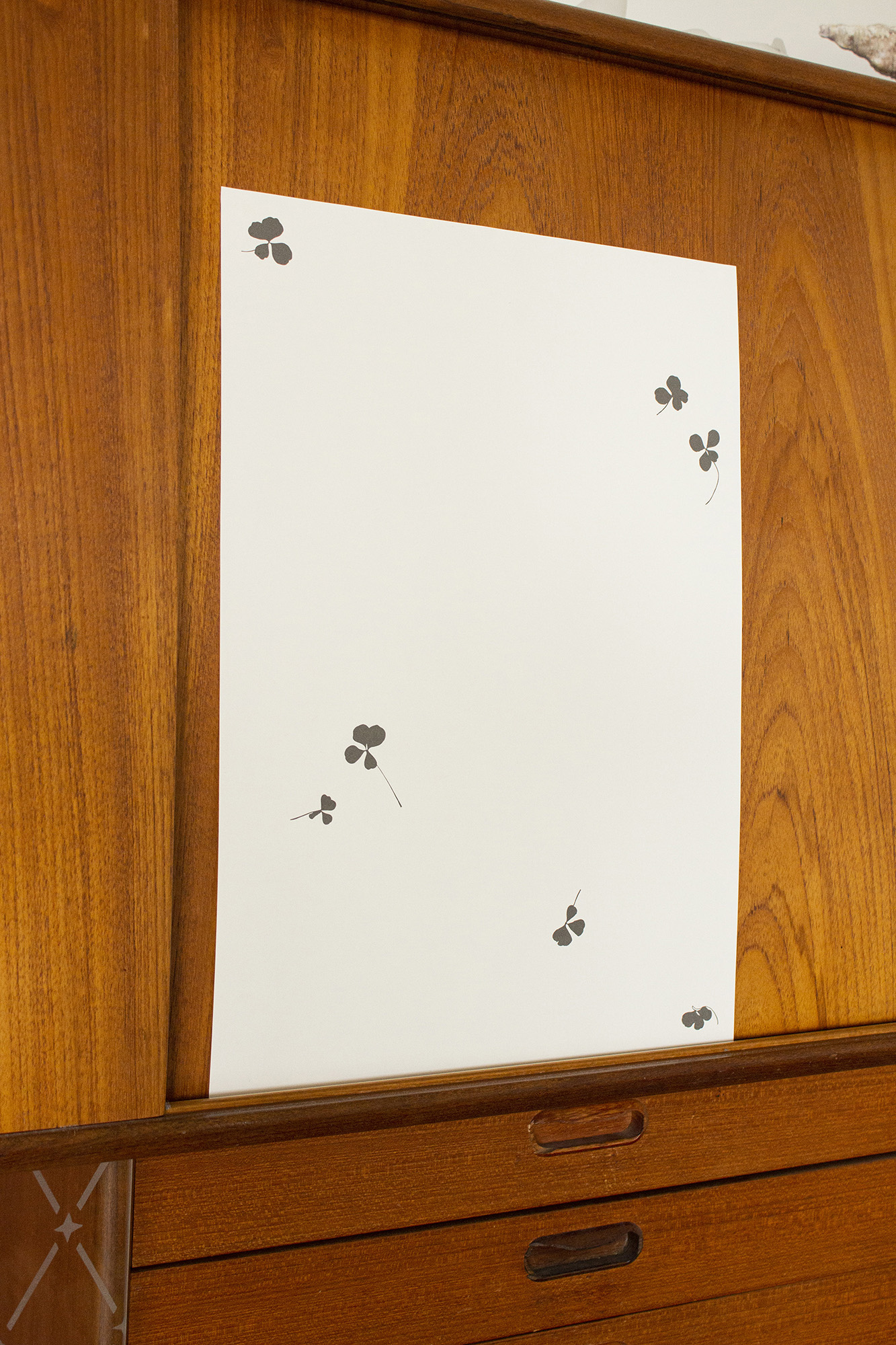 A piece of paper, speckled with dispersed four leaf clovers, leans up against the cabinet of a teak dresser.