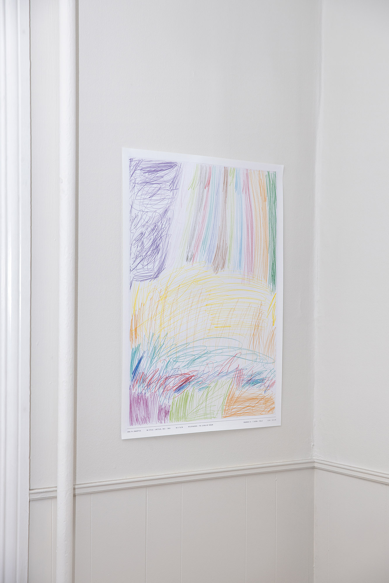 A poster hangs on a white wall in a corner of a room. A thin pipe is located close to the left side of the poster. The poster shows a colorful, childlike drawing, made with color pencils.